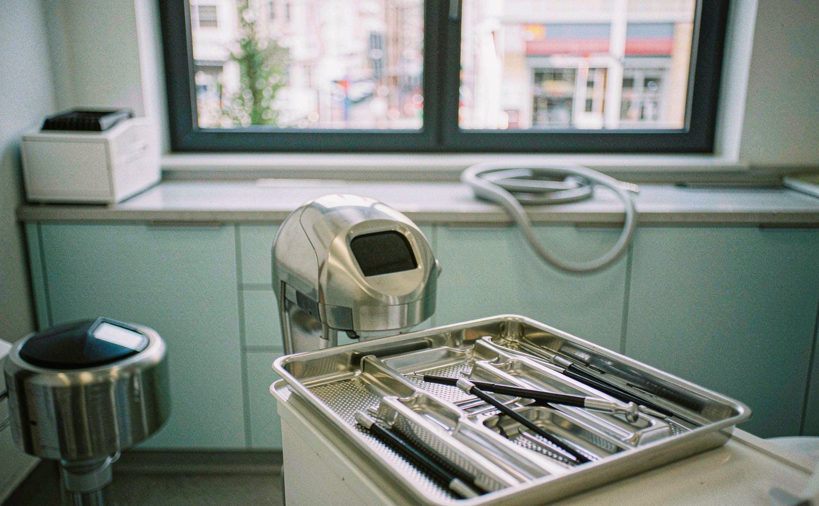 Stainless instrument tray holding slender dental instruments in a modern treatment room, with a grey hose coiled on the counter