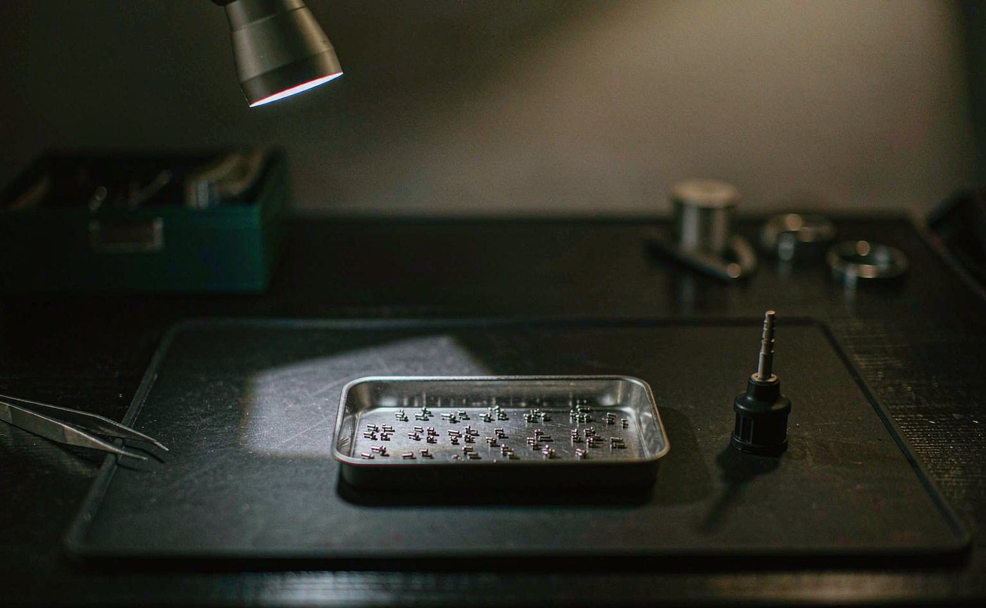 A precision repair workbench lit by a magnifier lamp, with miniature components in a metal parts tray